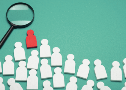Magnifying glass highlighting a red wooden figure among white figures, representing the process of hiring the right Medical Virtual Assistant for healthcare practices.